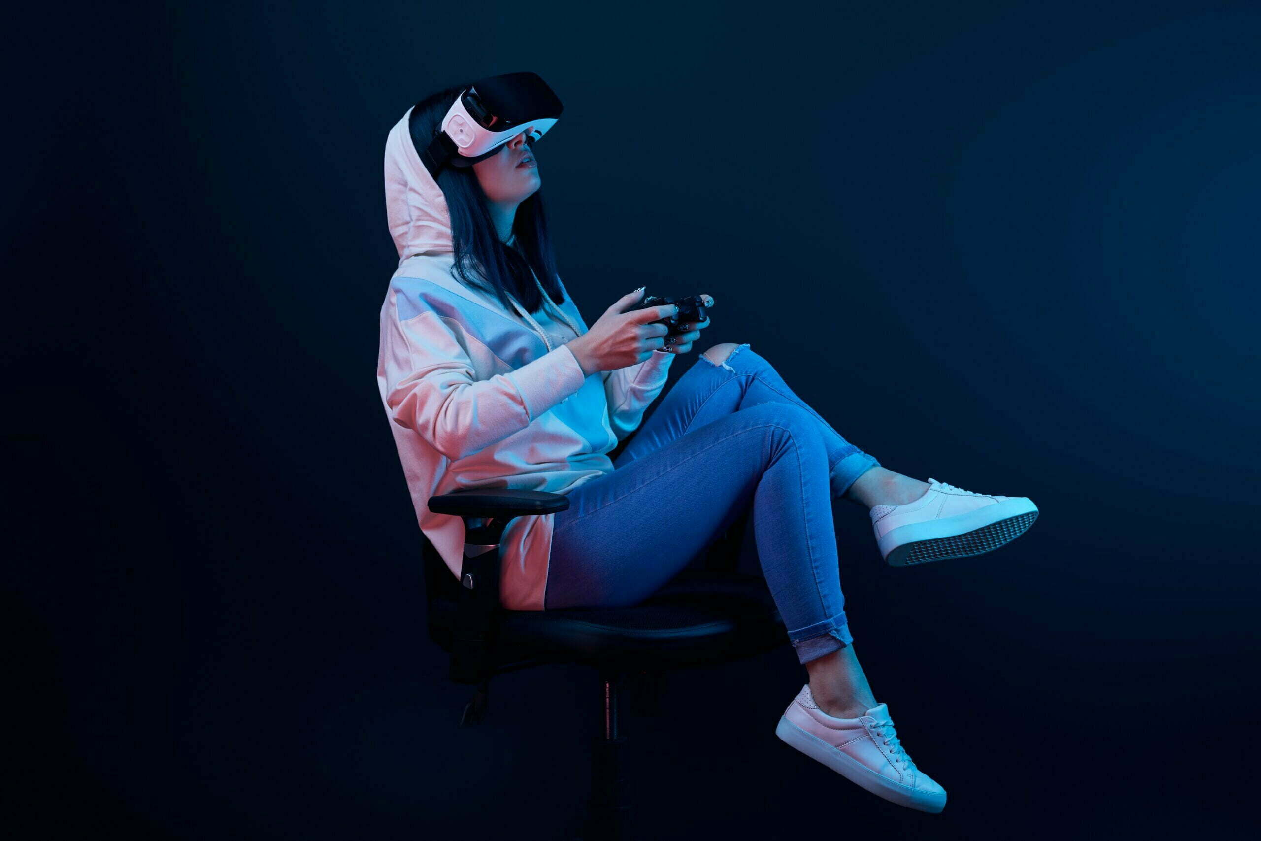 brunette woman sitting on chair and playing video game while wearing virtual reality headset on blue
